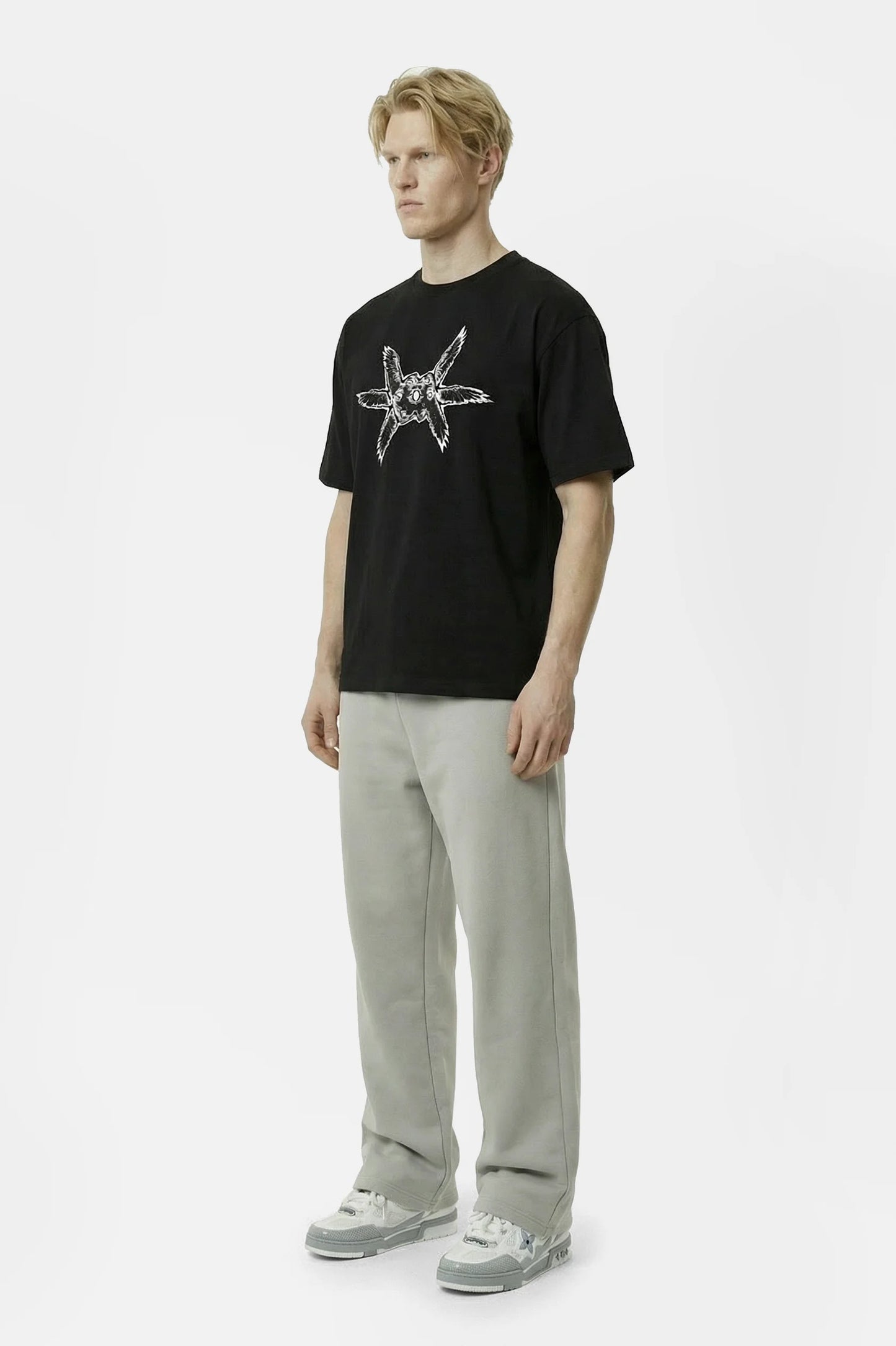 Man wearing a black oversized heavyweight t-shirt with a white ominous angel seraph seraphim design and light gray pants with light grey louis vuitton sneakers on a white background